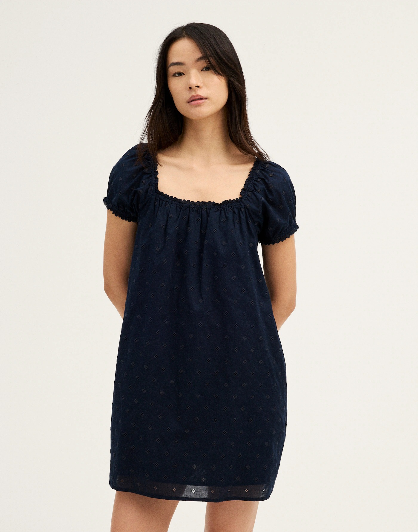 Person wearing a black eyelet-embroidered mini dress with a square neck and puff sleeves, standing with hands behind back