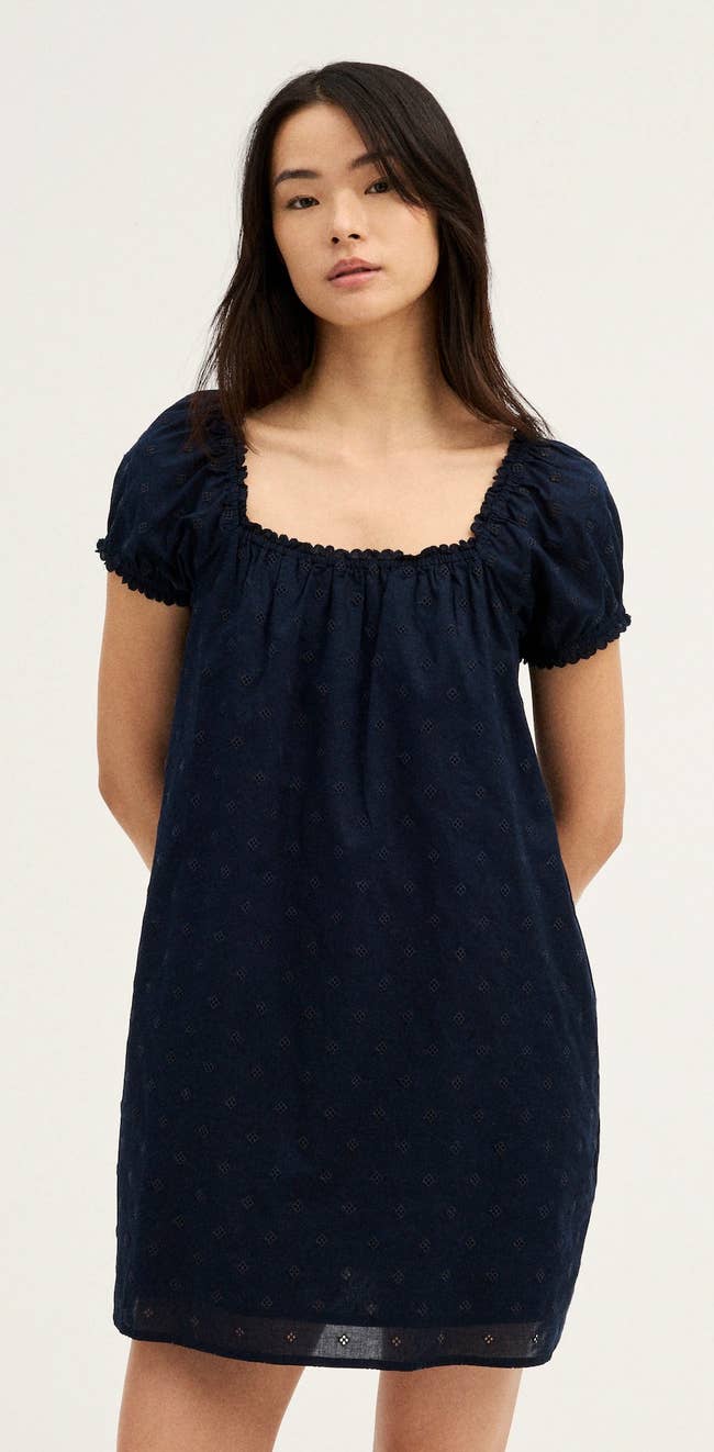 Person wearing a black eyelet-embroidered mini dress with a square neck and puff sleeves, standing with hands behind back