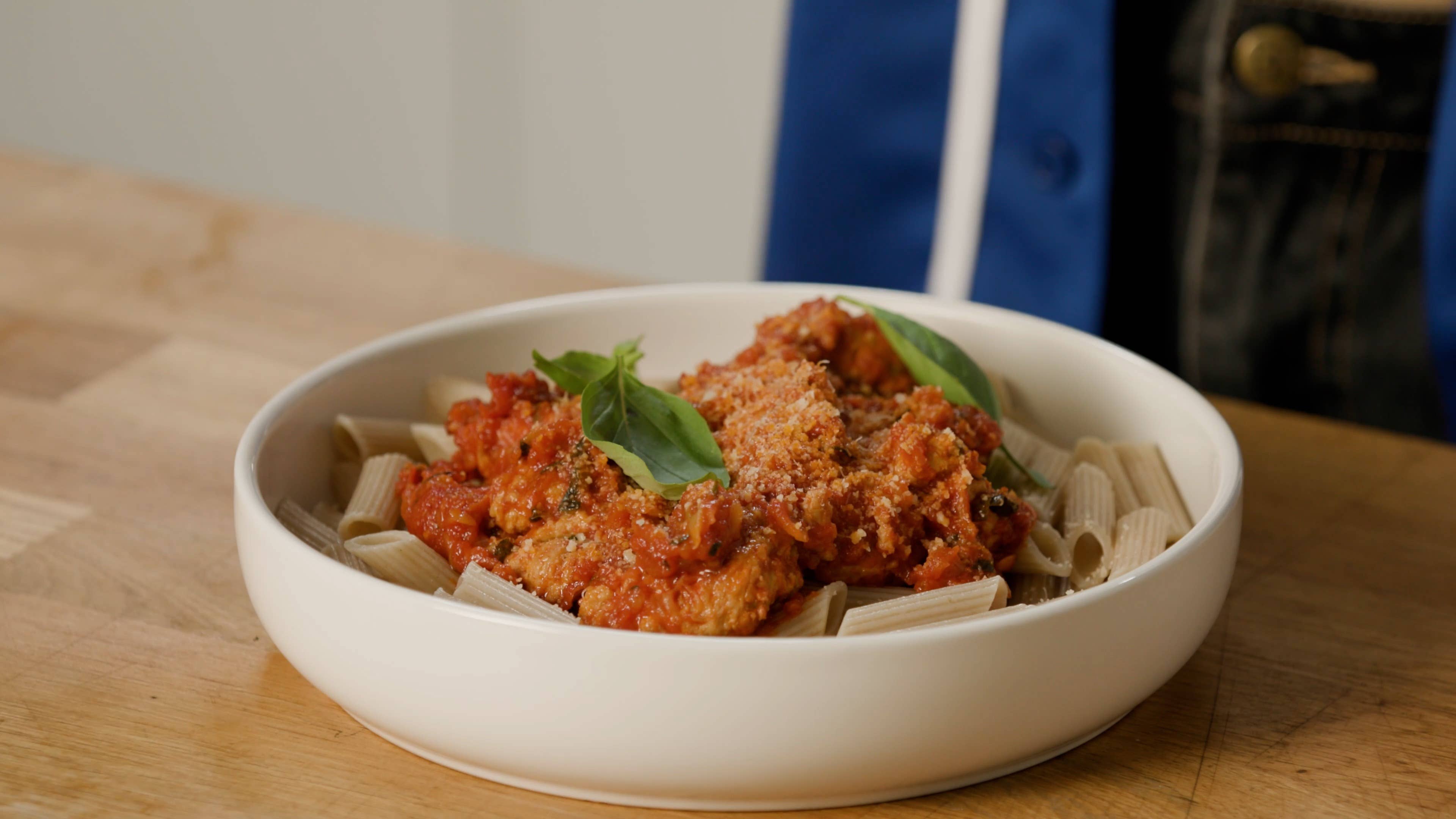 White bowl filled with pasta tube noodles, a red and meat sauce, and fresh basil, sitting on a wooden table.