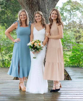 Three women outdoors, one in a bridal dress and two in sleeveless dresses, smiling, with a tree in the background