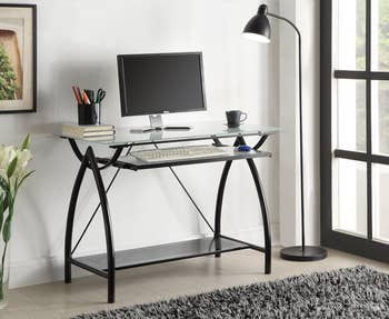 Modern glass-top computer desk with a monitor, keyboard, and office supplies, next to a floor lamp in a minimalist home office setting