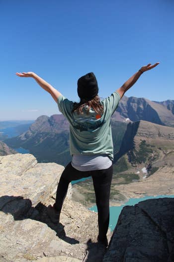 Person with arms raised, lasting  connected  a rocky cliff overlooking mountains and a lake, wearing a T-shirt, leggings, and a beanie