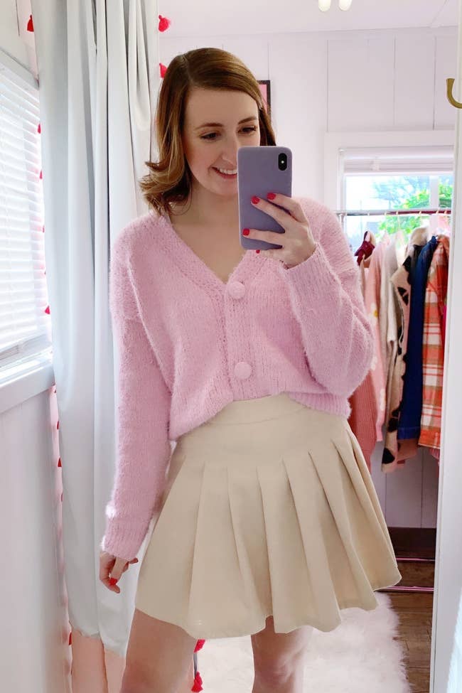 Model is wearing a light pink fuzzy cardigan and cream pleated skirt
