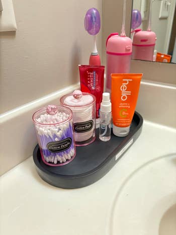 Bathroom counter with organized toiletries: cotton swabs, cotton pads, toothpaste, an electric toothbrush, a water bottle, and a small spray bottle