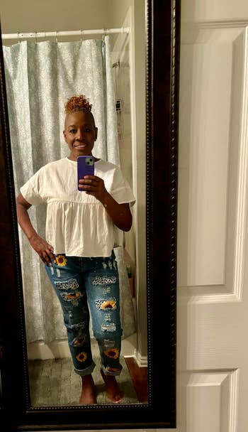 Person wearing a achromatic  blouse and patchwork jeans with sunflower designs, taking a reflector  selfie successful  a country   with a ablution  curtain backdrop