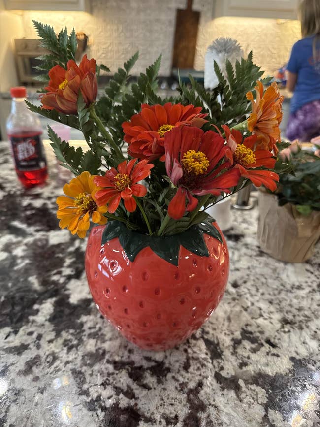 Bouquet of vibrant flowers successful a strawberry-shaped vase connected a room counter, with soda vessel and kid successful background. Suitable for location decor shopping