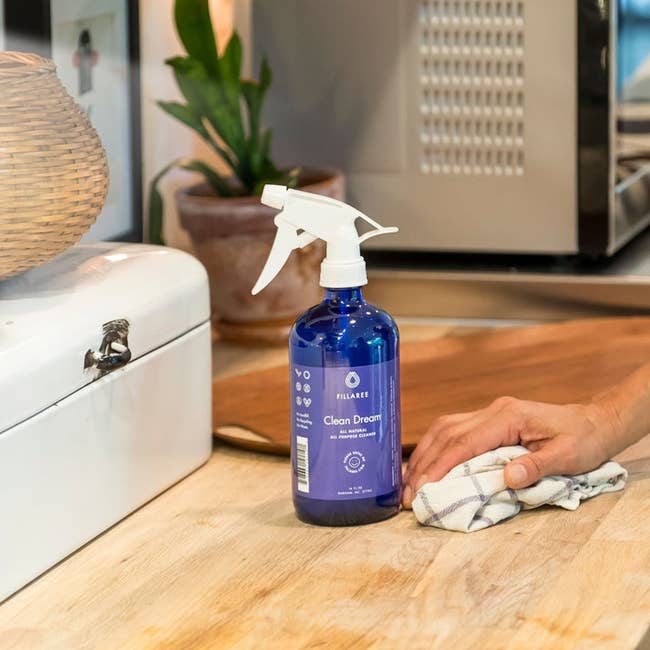 A person cleans a wooden countertop with a cloth next to a bottle of Fillaree Clean Dream spray in a kitchen setting