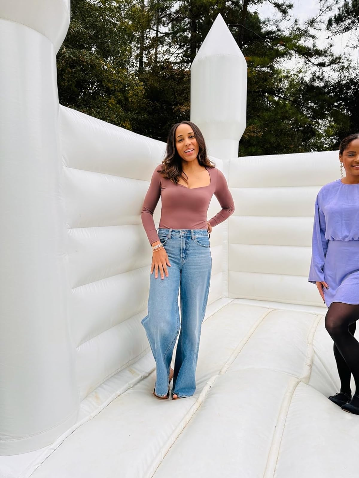 reviewer in a long-sleeve top and wide-leg jeans stands on an inflatable structure, smiling for a shopping article