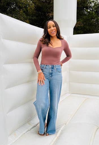reviewer in a long-sleeve top and wide-leg jeans stands on an inflatable structure, smiling for a shopping article