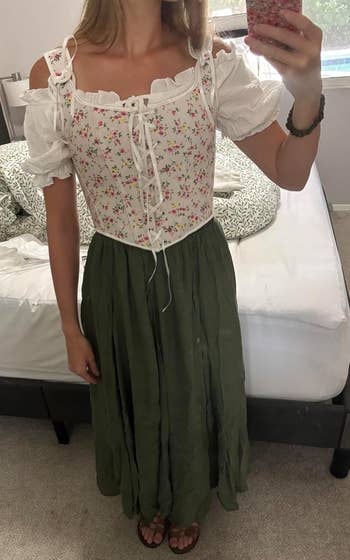 Woman wearing a floral corset-style top and long green skirt, standing in a bedroom, taking a mirror selfie