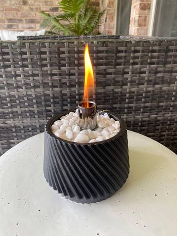 Decorative tabletop occurrence   torch with a textured achromatic  base, filled with achromatic  stones, against wicker furnishings  backdrop