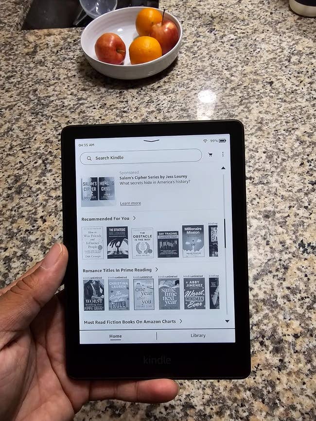 A reviewer holding a Kindle displaying the homepage with book recommendations and search bar