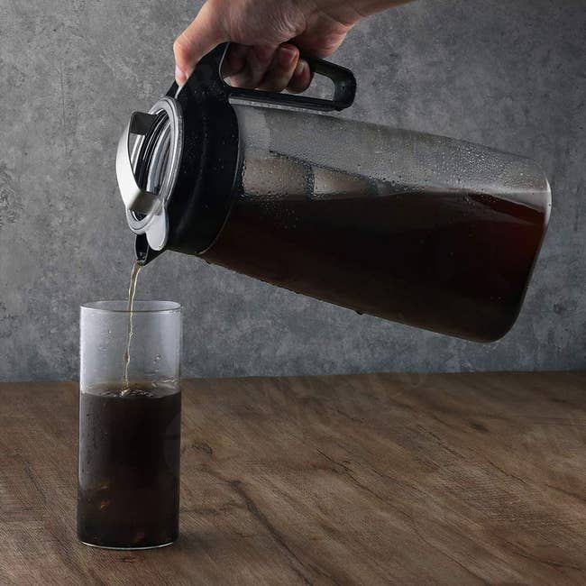 Clear container filled with cold brew and a filter pouring drink into a glass on a wooden table