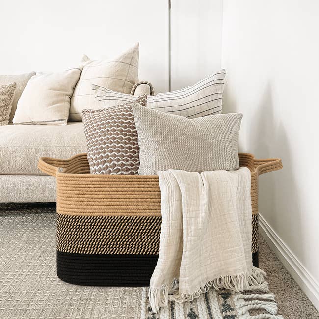 Woven handbasket  with handles holds a assortment  of textured propulsion  pillows and a light-colored blanket, adjacent  to a beige sofa connected  a patterned rug