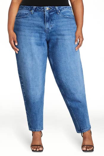 Person wearing high-waisted blue jeans and black heeled sandals, standing against a plain background