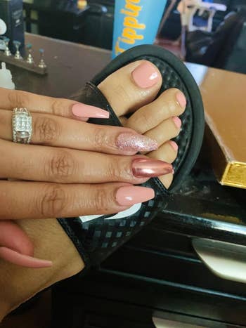 Manicured hand with a sparkly nail and a ring, resting on a foot with matching pedicure in a black sandal, on a desk