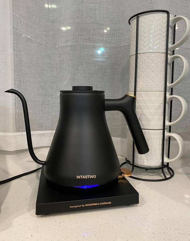 Electric kettle with a sleek, matte black finish on a countertop, next to a vertical stack of textured white mugs