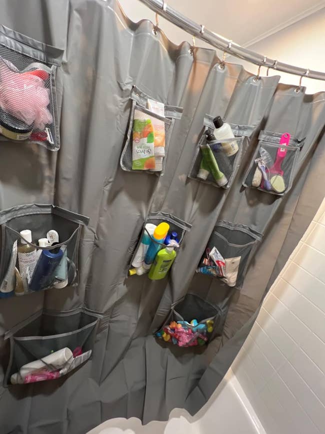 Reviewer's gray shower curtain liner with pockets filled with various bath products and toys, hanging in a bathtub