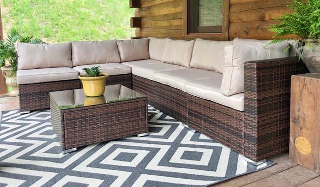 Outdoor patio set with beige cushions, wicker sectional sofa, glass-top wicker coffee table on geometric-patterned rug, and potted plants