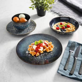 Waffles with strawberries and whipped cream, oatmeal with berries, and a vessel  of eggs are displayed connected  a eating  array  with cutlery and a plant