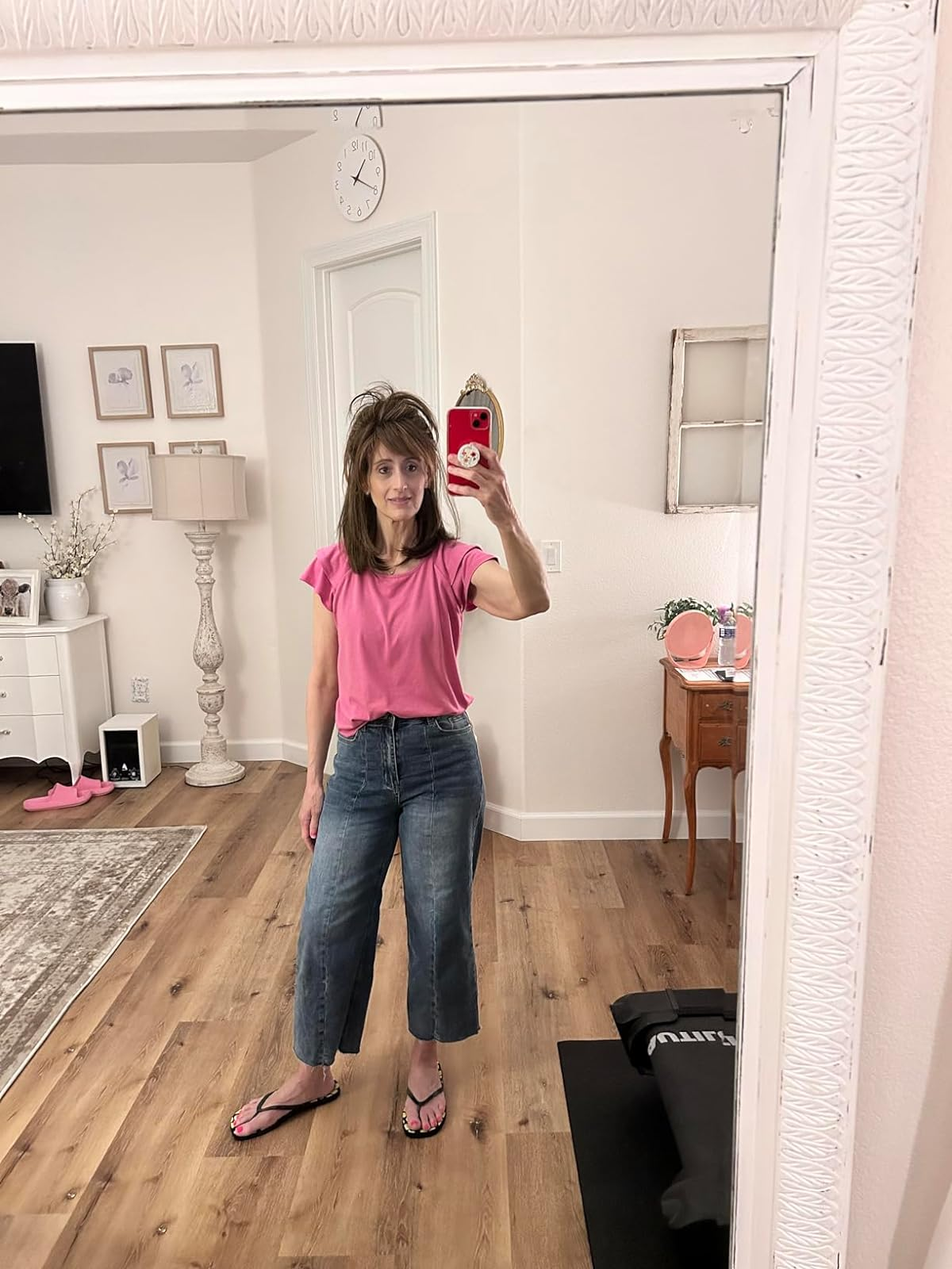 A reviewer taking a mirror selfie wearing a pink T-shirt, cropped jeans, and flip-flops in a home with decor and wooden floors