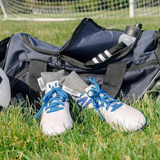A sports duffel bag with soccer gear, including cleats and a water bottle, rests on grass near a goal net. Ideal for athletic shopping articles