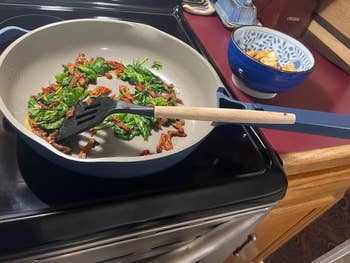 Frying pan on stove with spinach and sundried tomatoes; bowl of chopped ingredients nearby; kitchen setting for a meal prep concept