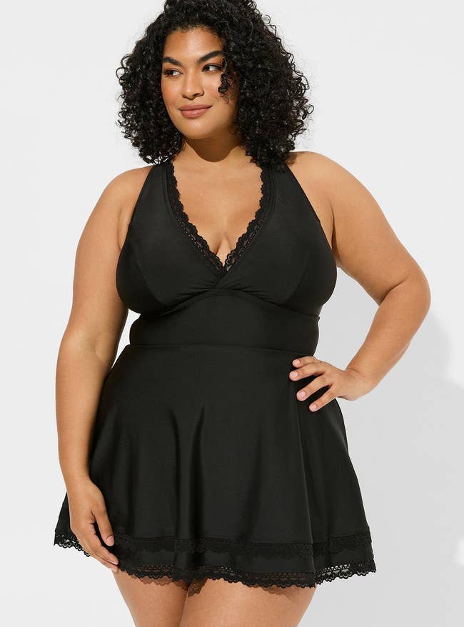 Woman in a black sleeveless swim dress with lace details, posing for a shopping-related article