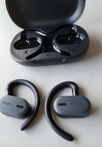 Reviewer's wireless TOZO earbuds with hooks next to open charging case 
