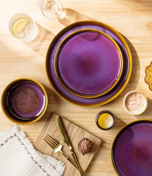Table mounting with circular purple dishes, a tiny bowl, golden utensils, a woody board, and glasses connected a woody table