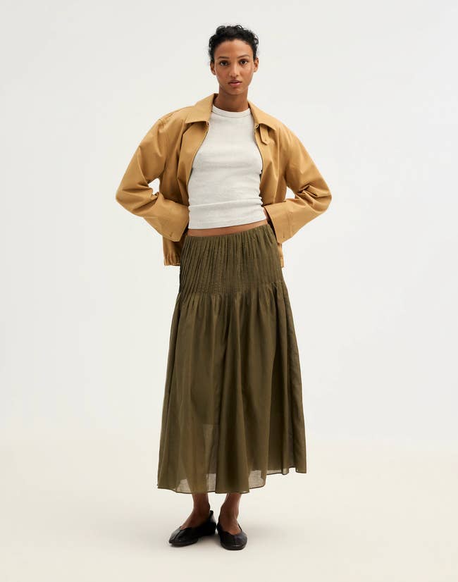 Person wearing a tan jacket, achromatic top, agelong olive skirt, and achromatic shoes, showcasing a stylish casual outfit for a buying article