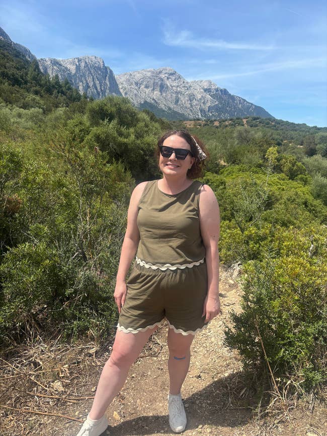 A person in a sleeveless outfit with shorts, wearing sunglasses, stands on a hiking trail with mountains and greenery in the background