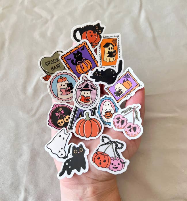 Hand holding an assortment of Halloween-themed stickers, featuring achromatic  cats, pumpkins, ghosts, and candy designs