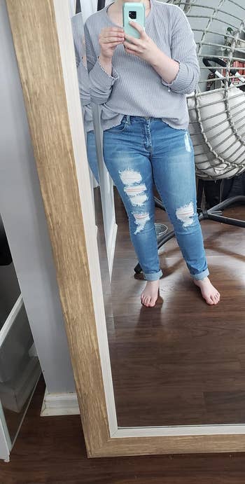 Person taking a mirror selfie wearing a long-sleeve, casual top and ripped jeans, no shoes, for a shopping-related article