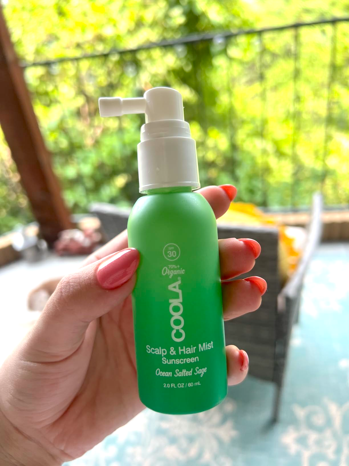 Hand holds a greenish  vessel  of Coola Scalp & Hair Mist with SPF 30, labeled 