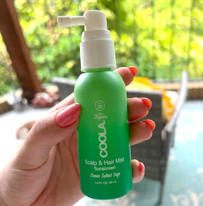 Hand holds a greenish vessel of Coola Scalp & Hair Mist with SPF 30, labeled