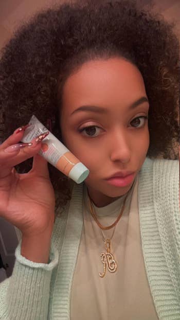 Person holding a skincare merchandise successful a tube, wearing a cozy textured cardigan and layered golden necklaces
