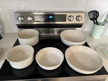 Five achromatic  pots and pans connected  a stovetop, acceptable   against a modern   room  backdrop with a sleek oven and utensil holder