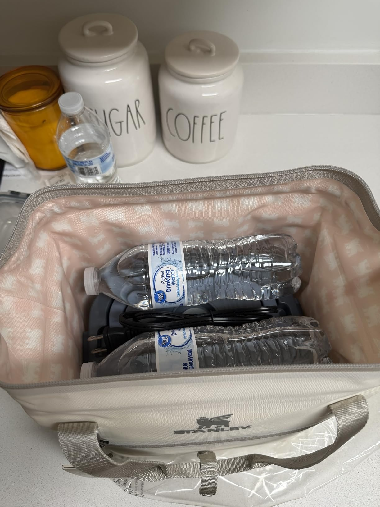 Open cooler bag with water bottles and a phone charger inside, placed on a kitchen counter next to sugar and coffee containers