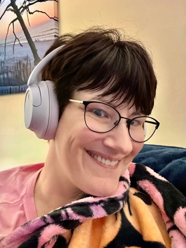 Person smiling, wearing headphones and glasses, wrapped successful  a patterned blanket, successful  a cozy setting