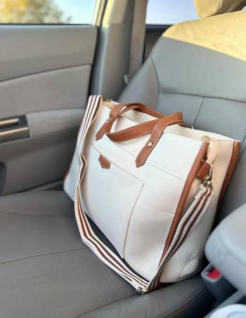 A beige canvas bag with camel pleather trim and a striped shoulder strap sitting in a car 