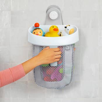 Hand placing bath toys into a white and gray mesh storage container mounted on a tiled wall.