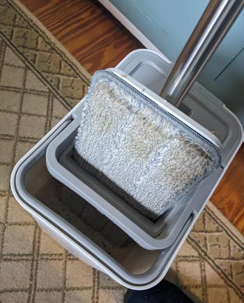 A gray flat mop vertically pushed into a rectangular shaped mop bucket 