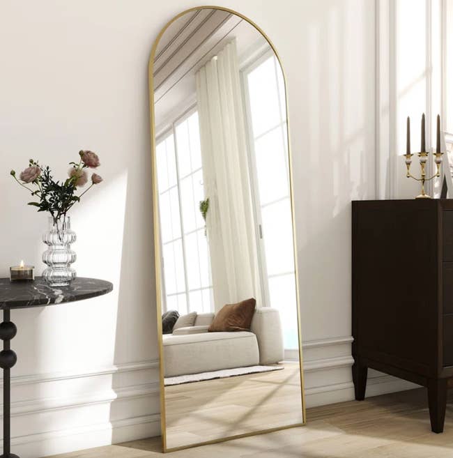 WordPress 網站 A modern living room features a tall, arched floor mirror with a gold frame, reflecting a white couch with brown pillows, accompanied by a dark wooden dresser and a side table with a vase of flowers