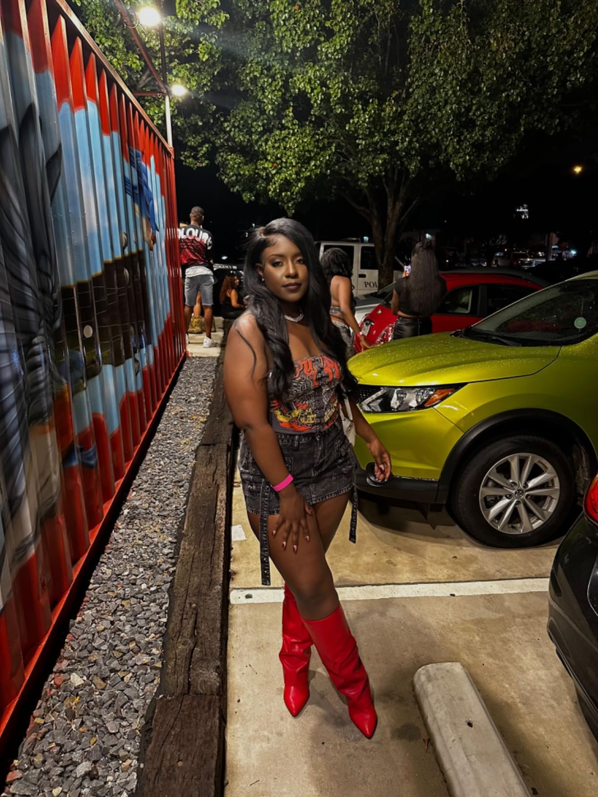 Person in graphic tee, black skirt, and red boots stands outdoors near a mural and green car, at night, with people in the background