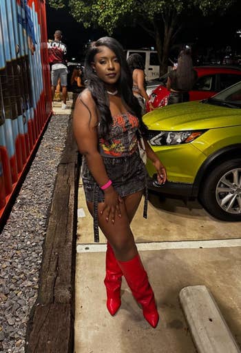 Person in graphic tee, black skirt, and red boots stands outdoors near a mural and green car, at night, with people in the background