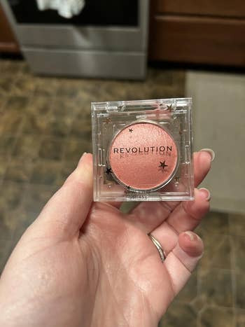 Hand holding a Revolution blush compact with a shimmery peach shade, shown successful a room setting
