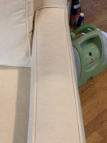 A half-view of a beige upholstered chair arm showing a vertical stain and a green Bissell Little Green ProHeat cleaner on a wooden floor