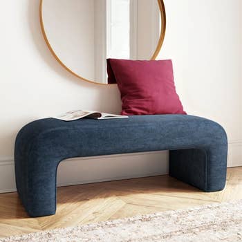 Blue curved bench with a red pillow, open magazine, and a round mirror above, placed in a stylishly decorated interior