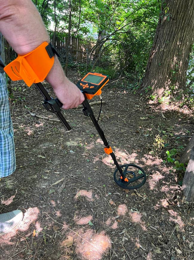 Person using an orange metal detector in a wooded area, focused on search